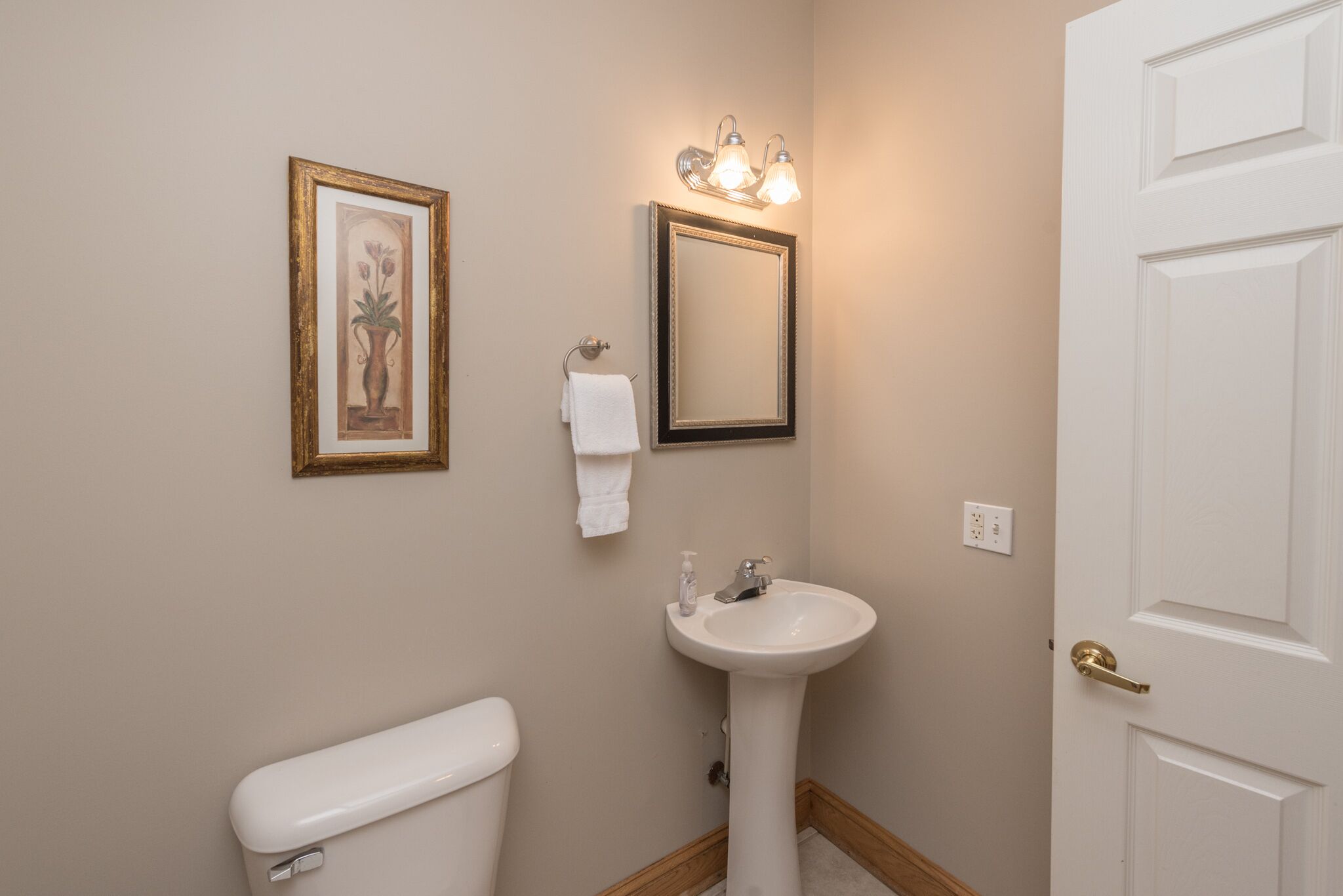 A bathroom with a toilet, sink, framed artwork, and a mirror with lights.