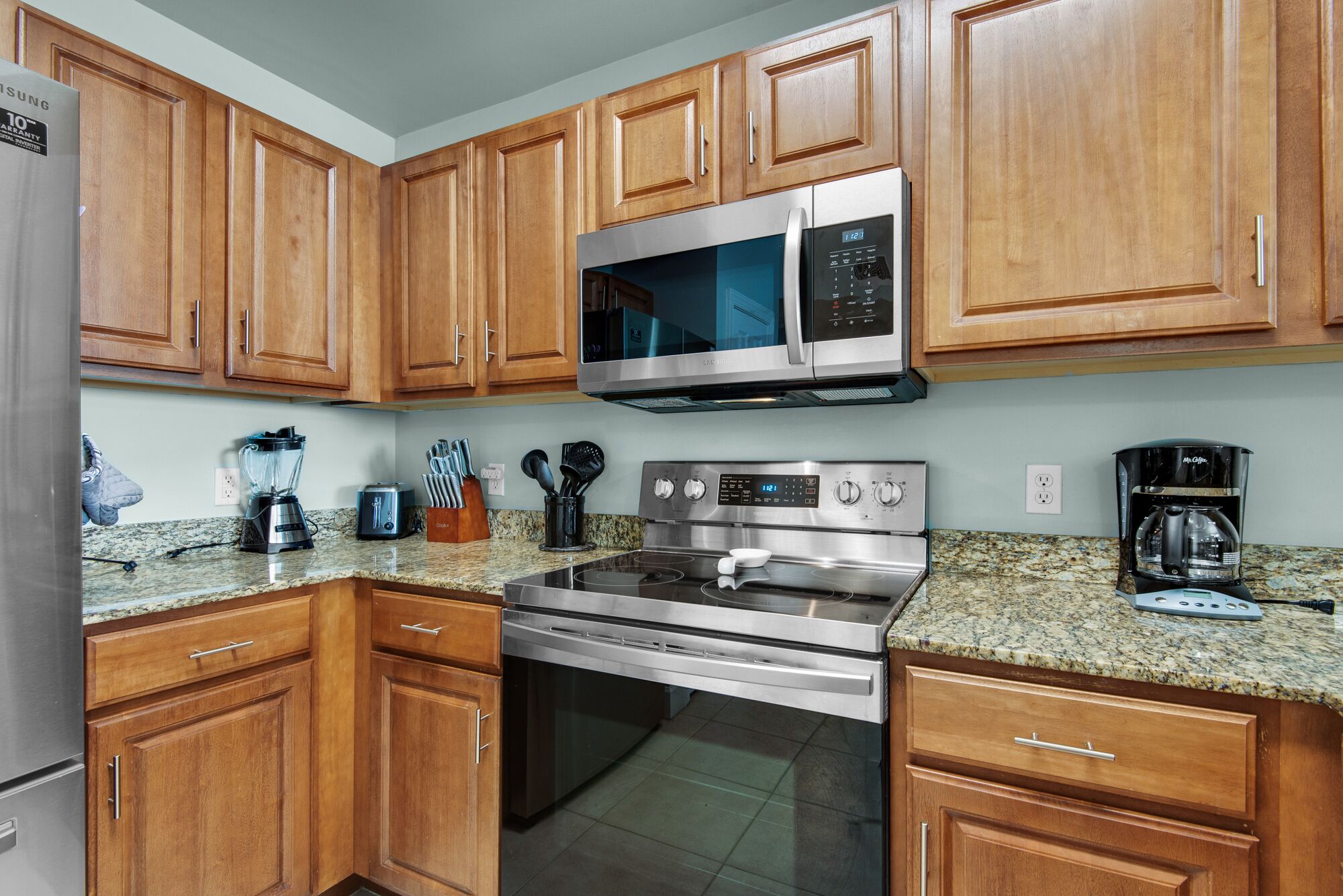 A kitchen with wooden cabinets, stainless steel appliances including a microwave, stove, and refrigerator, granite countertops, and various kitchen utensils.