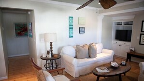 A living room with a white sofa, a wooden coffee table, and a wicker chair. There is a lamp on a side table, and a television in the background. - Gulf Winds Down 3/2 in Grayton, comm.pool, short walk to beach (Santa Rosa Beach)