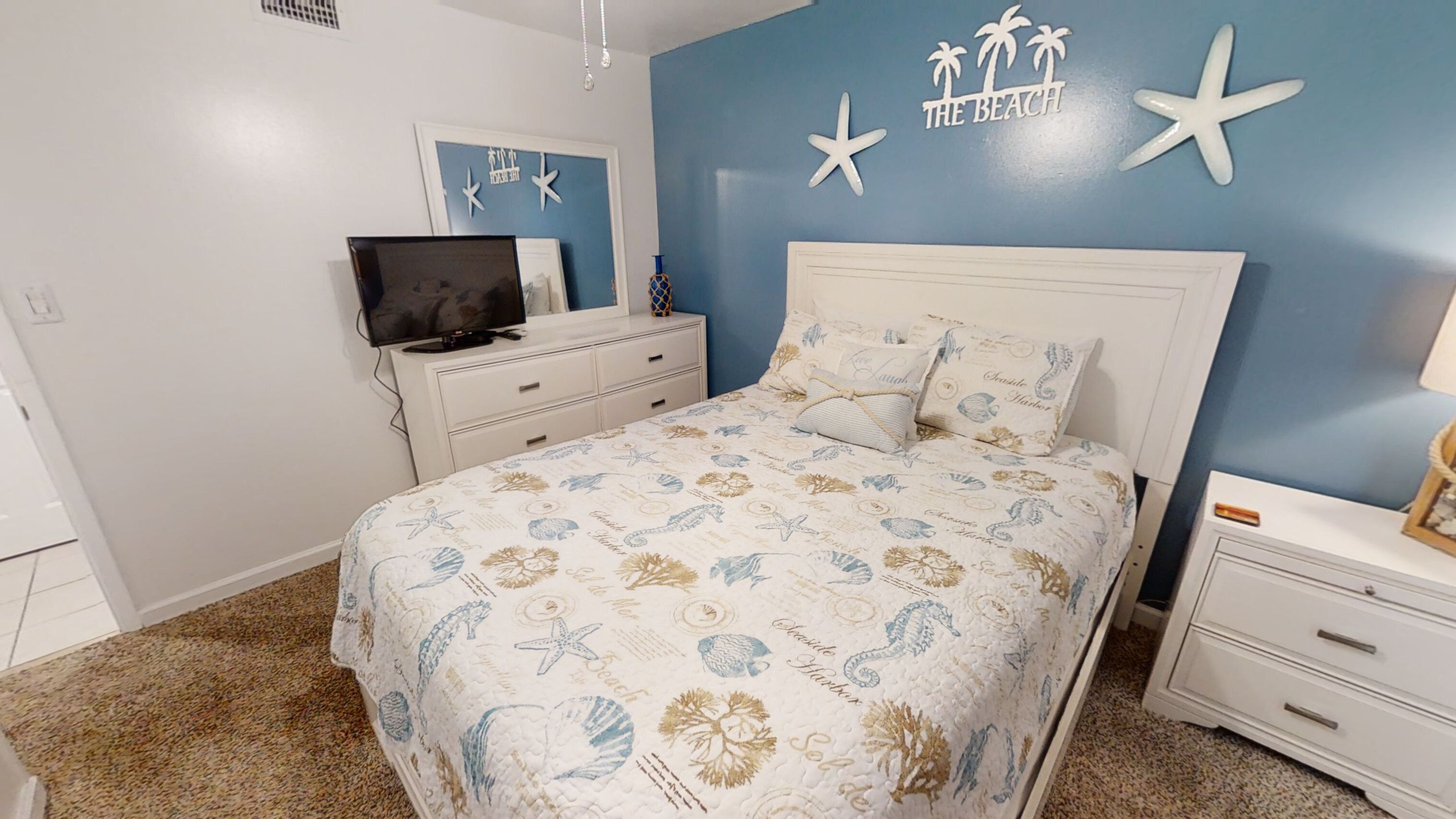 A bedroom with a bed, a TV on a dresser, and a wall with a beach theme.