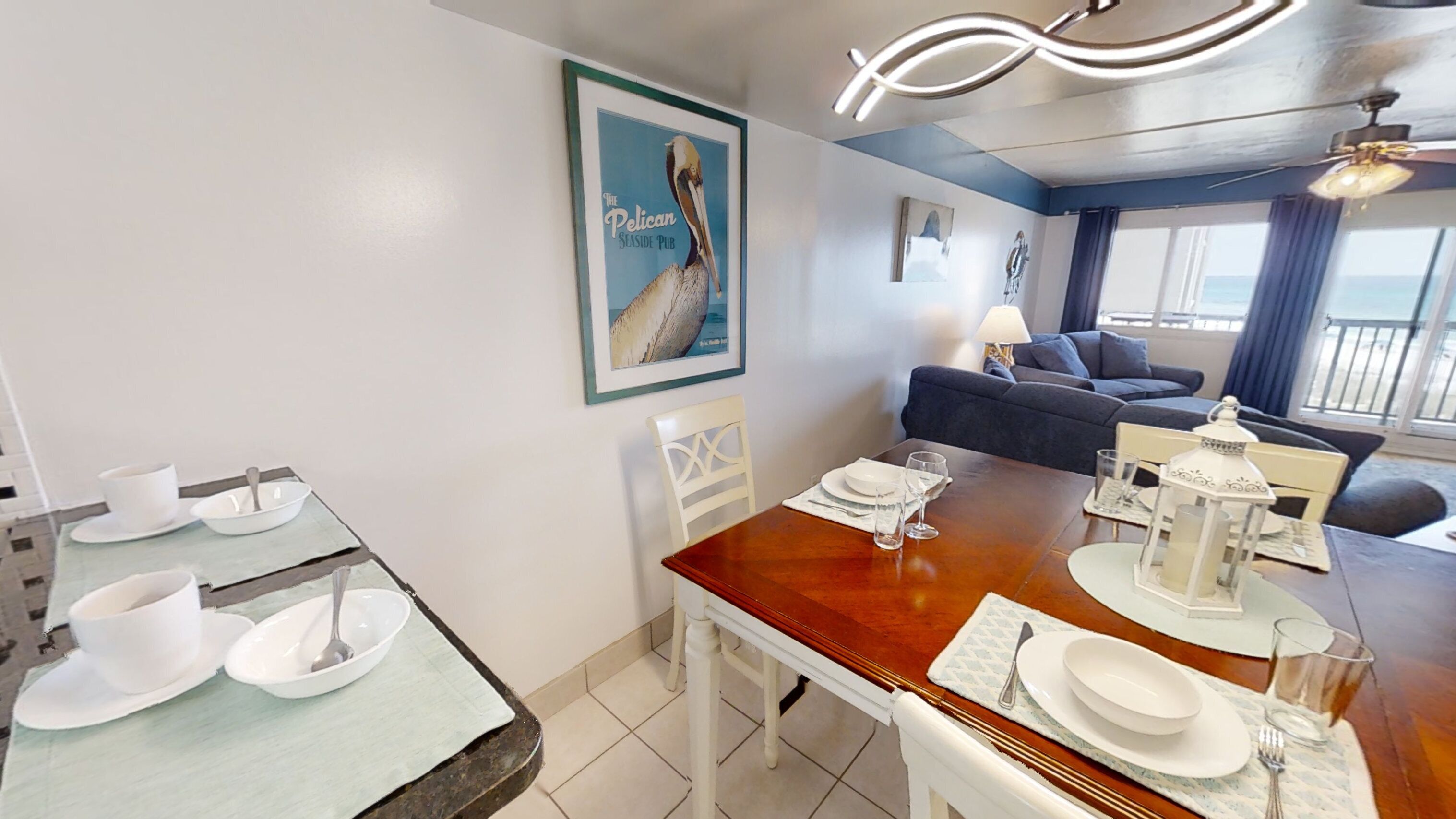 Un coin repas avec une table en bois dressée pour quatre personnes, des chaises blanches et un tableau encadré représentant un pélican, accroché au mur.