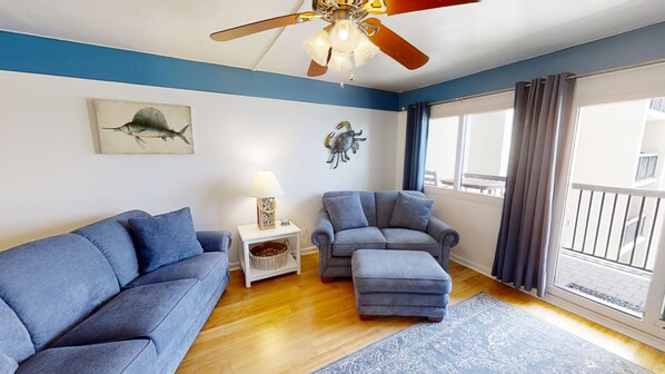 Una sala de estar con un sofá azul, un sillón gris, una mesita con una lámpara y un ventilador de techo.