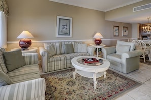 A living room with a striped sofa, a round coffee table, and two armchairs. There are two lamps on side tables, a framed picture on the wall, and a patterned rug on the floor.