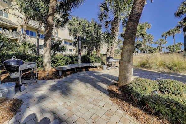 A paved outdoor area with a barbecue setup, benches, and palm trees.