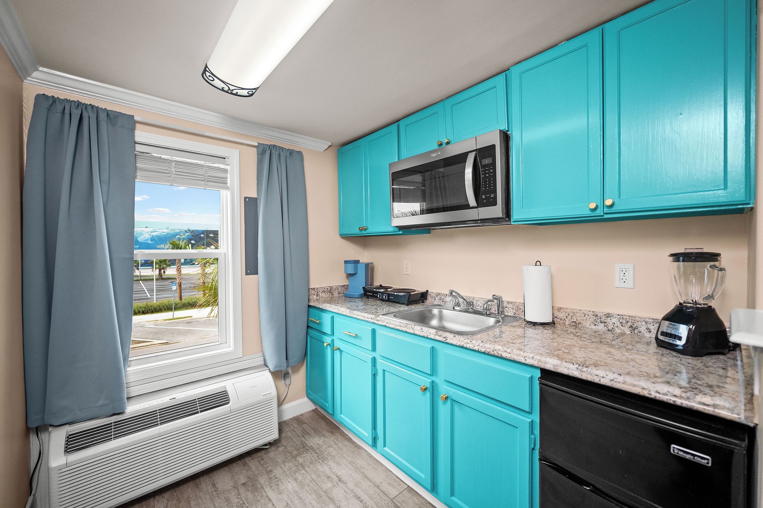 A kitchen with teal cabinets, a microwave, a sink, a blender, and a window with curtains.