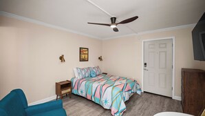 Un dormitorio con una cama, un sillón azul, una mesita de noche blanca, un ventilador de techo y una puerta.