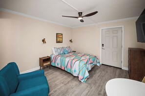 A bedroom with a bed, a blue armchair, a white bedside table, a ceiling fan, and a door. - Gulf Shores Condo for Two | Walk-to-Beach Location (Gulf Shores)