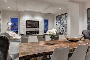 Un salon moderne avec un mur d’accent en pierre, une cheminée, un grand tableau abstrait et un coin repas comprenant une table en bois et des chaises blanches.