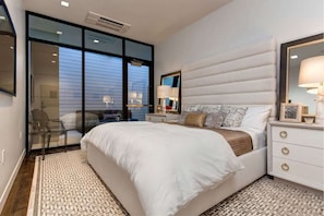 Une chambre avec un grand lit, une table de chevet avec une lampe, un miroir et une porte-fenêtre coulissante donnant sur un balcon.