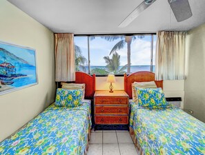 A hotel room with two single beds, a wooden dresser, a nightstand with a lamp, and a large window offering a view of palm trees and the ocean.