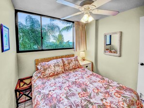 Ein Schlafzimmer mit Bett, Nachttisch, Spiegel und Fenster mit Blick auf Palmen.