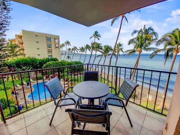 A balcony with a table and chairs, overlooking a pool and a beach with palm trees.