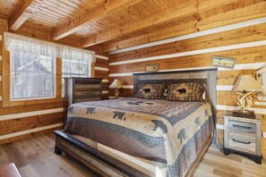 A log cabin bedroom with a large bed, wooden walls, a window with blinds, and a nightstand with a lamp.