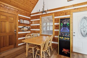 A wooden interior with a dining table and chairs, a Pac-Man arcade machine, and a white door.