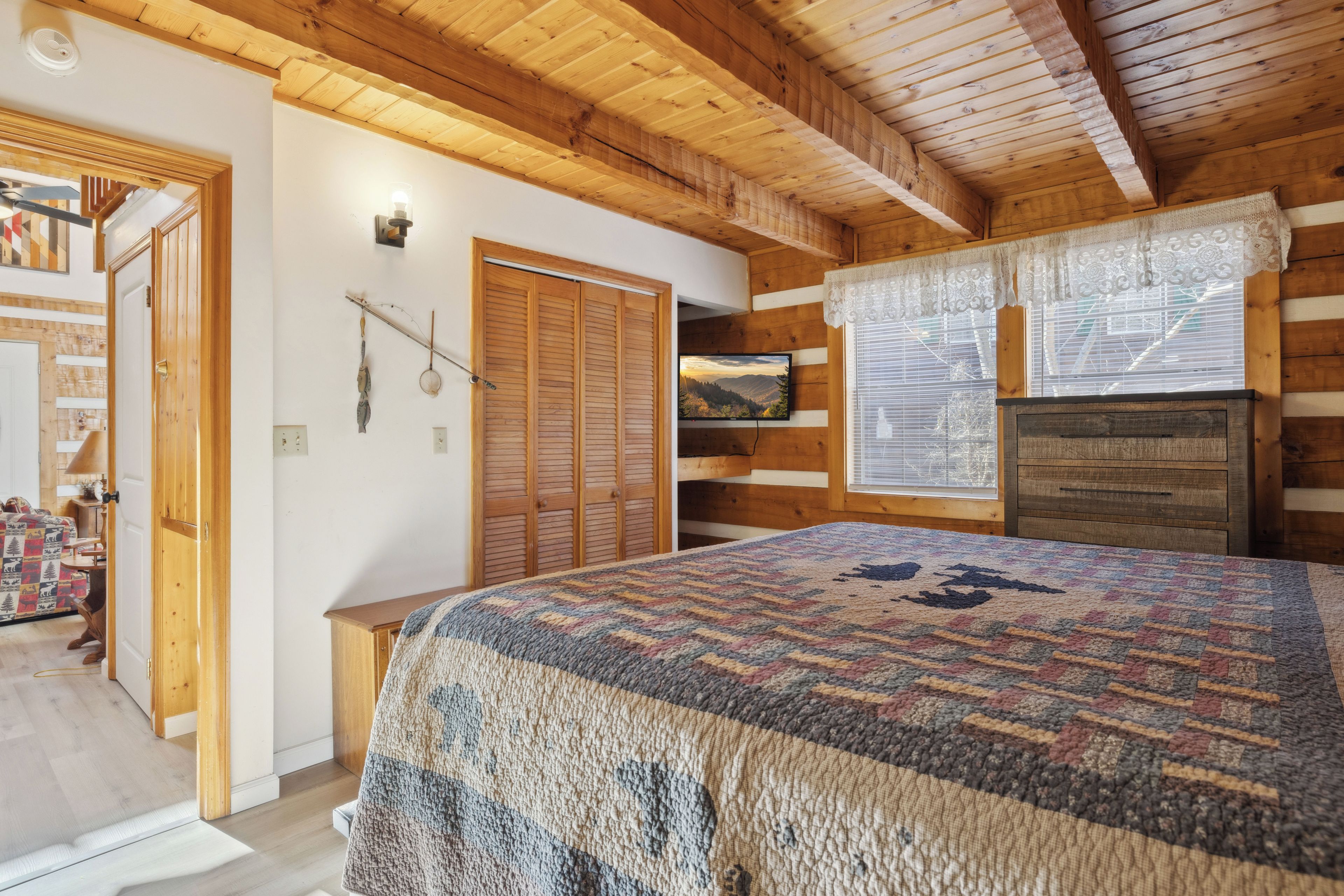 Une pièce chaleureuse avec un plafond en bois, un lit recouvert d’une couverture à motifs, un téléviseur et une fenêtre ornée de rideaux en dentelle.