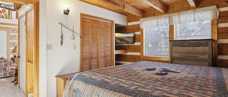 Una habitación acogedora con techo de madera, una cama con manta estampada, televisión y ventana con cortinas de encaje.
