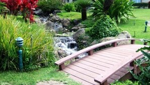 A wooden bridge over a small stream in a garden with tropical plants and a building in the background. - Palms at Wailea #205 Fully Remodeled, Ground Floor Unit, Shared Hot Tub, A/C (Wailea)
