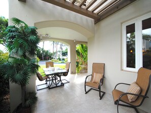 A covered patio area with a table and chairs, surrounded by greenery and a view of the outdoors.