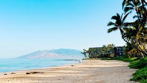 A beach with a clear view of the ocean and a mountain in the distance. There is a building with multiple floors and palm trees on the right side. - Palms at Wailea #205 Fully Remodeled, Ground Floor Unit, Shared Hot Tub, A/C (Wailea)