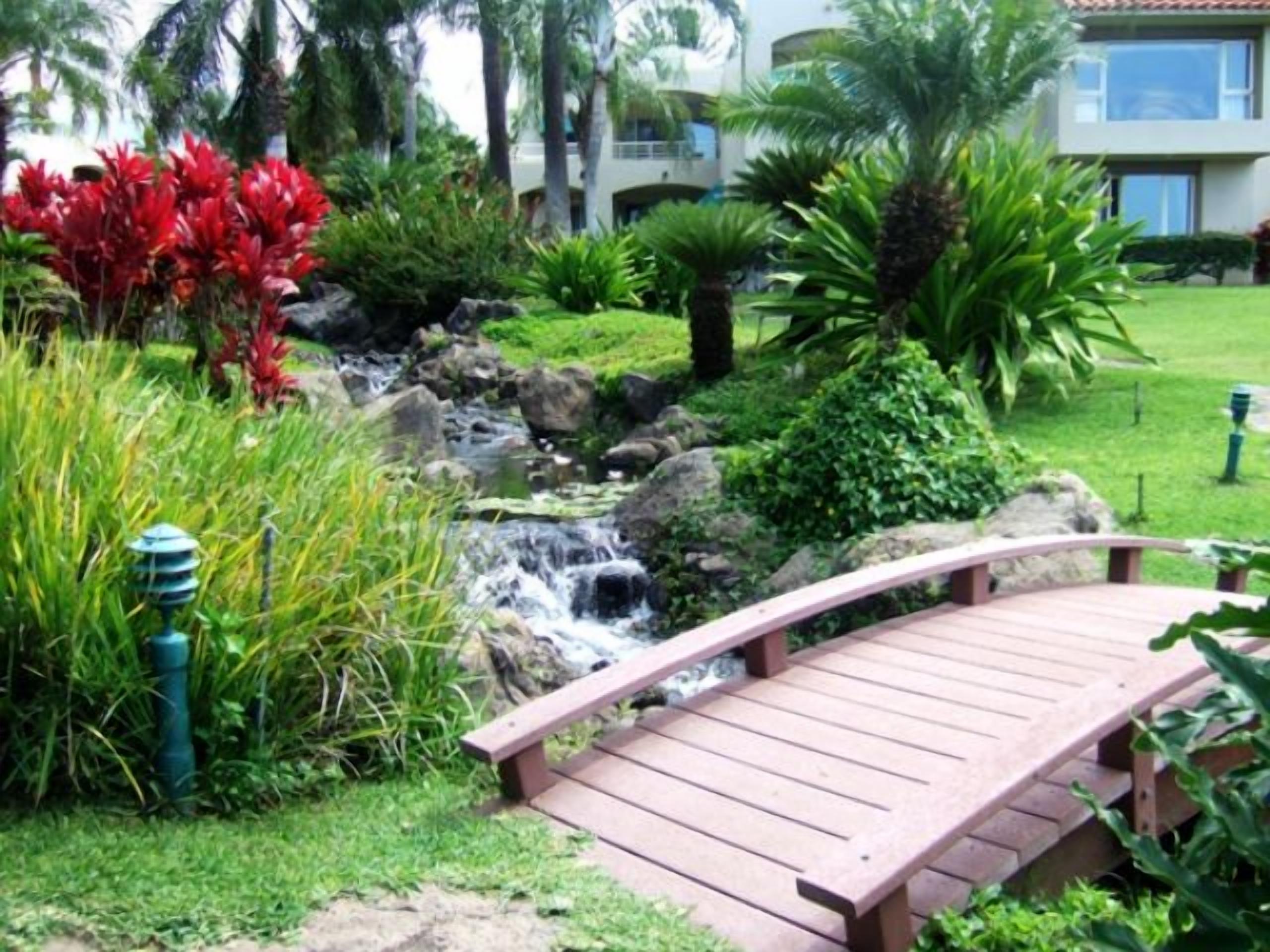 A wooden bridge over a small stream in a garden with tropical plants and a building in the background.