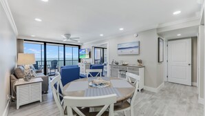 A spacious living area with a dining table, blue chairs, a sofa, and a balcony with a view. - SUNDIAL A306 - Direct Beachfront Sanibel Condo (Sanibel)