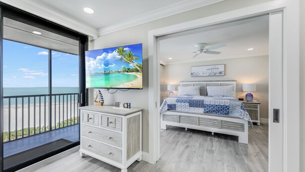 A bedroom with a large window overlooking a beach, a bed with a blue and white quilt, a white dresser, and a ceiling fan. - SUNDIAL A306 - Direct Beachfront Sanibel Condo (Sanibel)