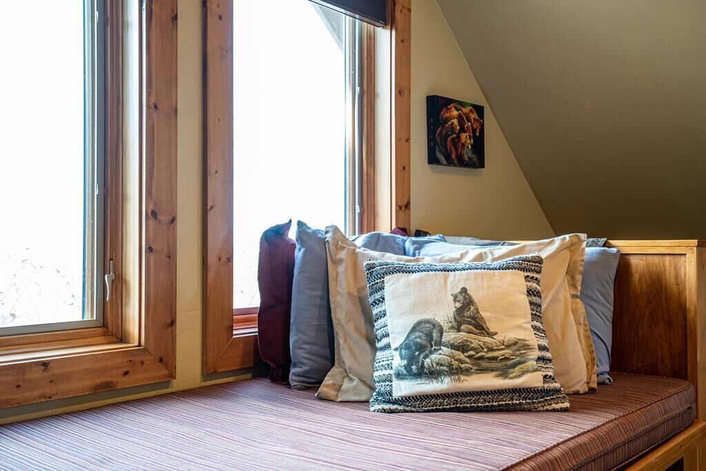 Ein gemütliches Zimmer mit schrägen Wänden, Holzbalken, einem Fenster mit Jalousien, einer gepolsterten Bank mit Kissen und einem Gemälde an der Wand.