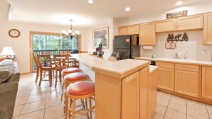 A modern kitchen with wooden cabinets, a large island with stools, a refrigerator, and a dining area with chairs and a table. - Chimney Ridge Sunflower Townhome: Walk to Lift, Private Hot Tub (Breckenridge)