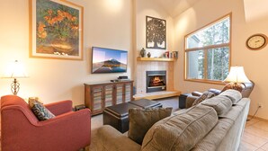 A living room with a sofa, armchair, and a fireplace. There is a television and a bookshelf. - Chimney Ridge Sunflower Townhome: Walk to Lift, Private Hot Tub (Breckenridge)