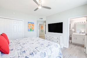 Une chambre à coucher comprenant un lit, un ventilateur de plafond, une télévision, une coiffeuse avec un miroir et une porte donnant sur une autre pièce.