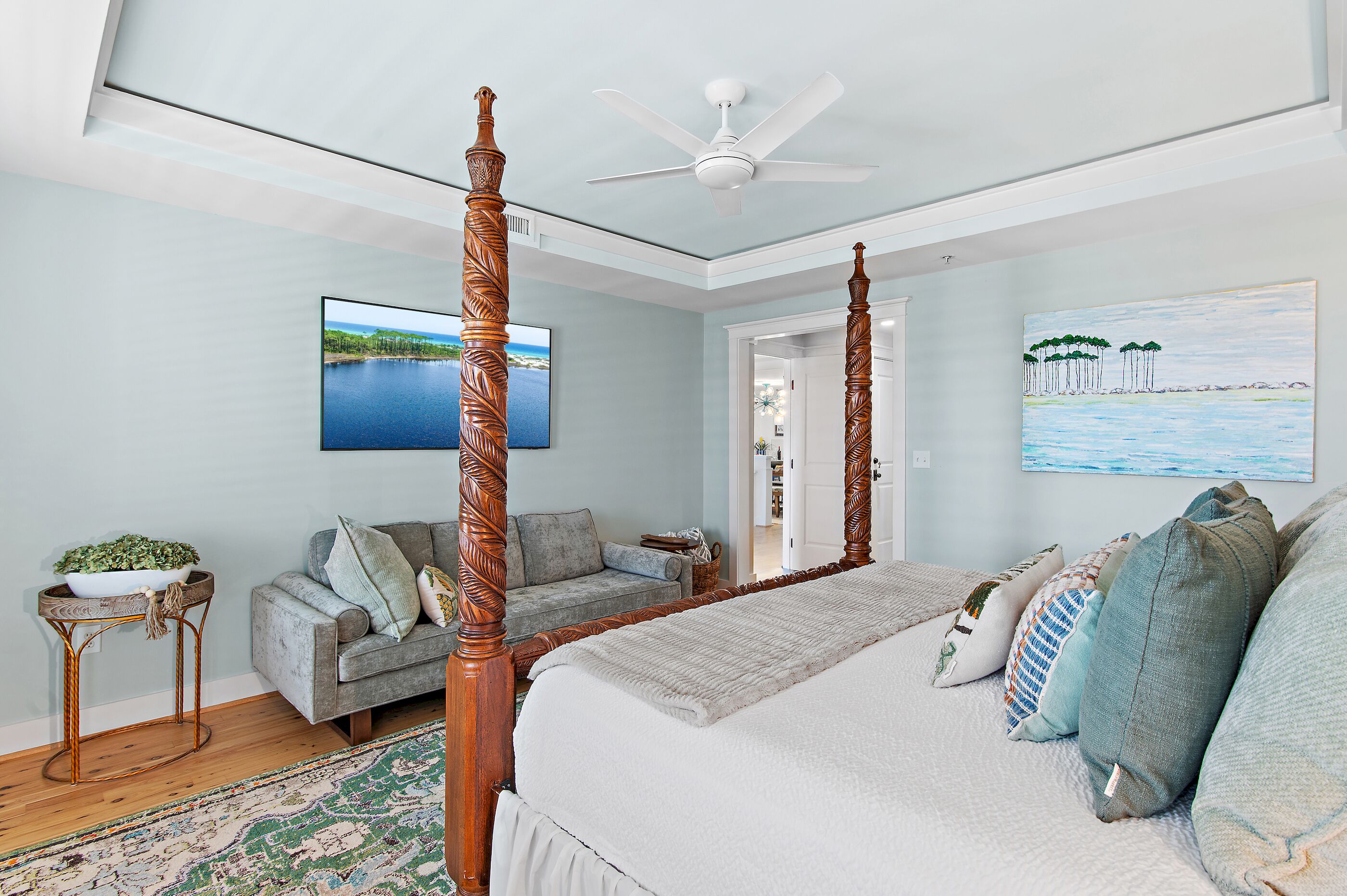 A bedroom with a four-poster bed, a ceiling fan, a television, and a painting of a beach scene.