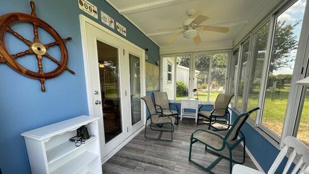 A covered porch with a ceiling fan, a ship's wheel decoration, and a view of the backyard through large windows. - Relaxation & Privacy w/Golf Cart Conveniently Located NOW Waiting for YOU! (The Villages)