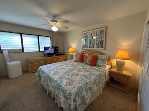 A bedroom with a bed, a ceiling fan, a TV, a window with blinds, and a painting on the wall. - Waiohuli Beach Hale #C-210 Oceanfront View, A/C Throughout Unit, Shared Pool (Kihei)