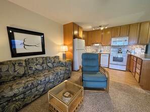 A living room with a sofa, a chair, a coffee table, and a kitchen area with wooden cabinets, a refrigerator, and a dishwasher.