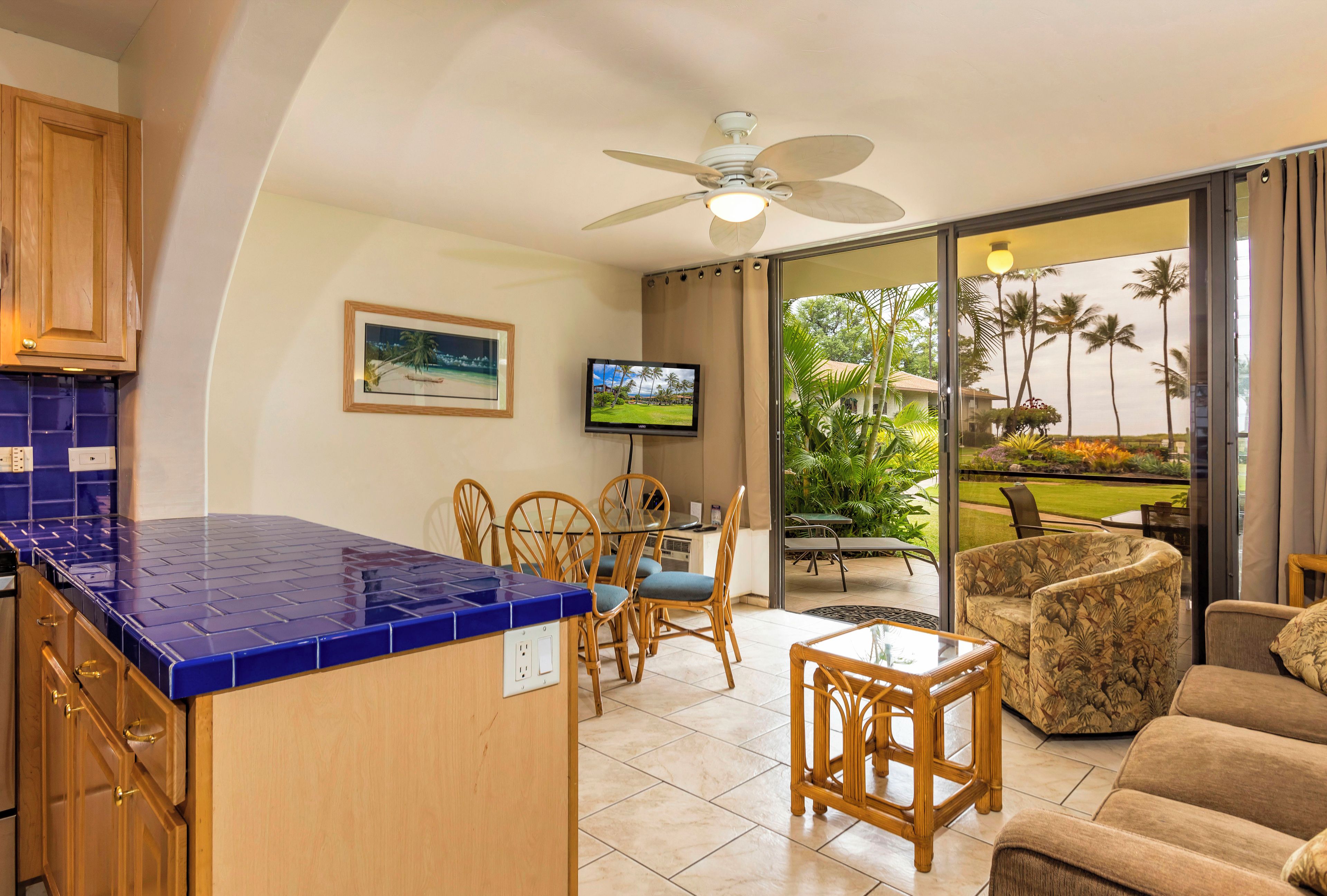 A kitchen with blue tiles, a dining area with wooden chairs, a television, and a balcony with a view of palm trees.