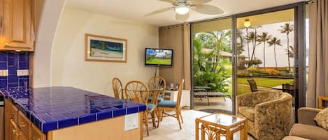 A kitchen with blue tiles, a dining area with wooden chairs, a television, and a balcony with a view of palm trees.