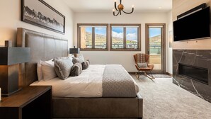 A bedroom with a large bed, a TV, a fireplace, and a view of the outdoors.
