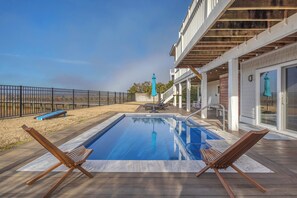 A swimming pool with two wooden lounge chairs, a covered patio area, and a view of the beach. - Sweet Shirl. A Spacious Sound Front Home with Private Pool! (Wrightsville Beach)