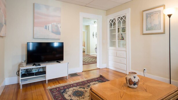 Een woonkamer met een flatscreentelevisie, een witte televisiemeubel, een houten bijzettafeltje en een kamerbreed tapijt.