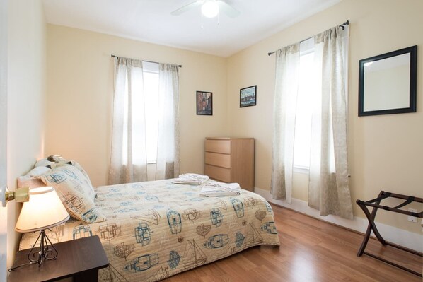 A bedroom with a bed, a nightstand, a dresser, and two windows with curtains.