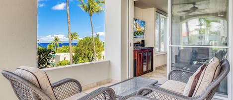 A balcony with wicker furniture, a glass table, and a view of the ocean and palm trees.