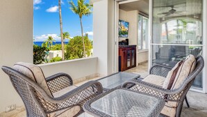 A balcony with wicker furniture, a glass table, and a view of the ocean and palm trees. - Palms at Wailea #1105 Lovely Ocean View, Near Complex Pool and Shared Hot Tub (Wailea)