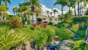 A lush garden with a small stream, palm trees, and a building with balconies. - Palms at Wailea #1105 Lovely Ocean View, Near Complex Pool and Shared Hot Tub (Wailea)