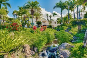 Un rigoglioso giardino con un piccolo ruscello, palme e un edificio con balconi.