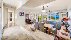 A spacious living area with a dining table, wicker chairs, a sofa, and a kitchen in the background. - Palms at Wailea #1105 Lovely Ocean View, Near Complex Pool and Shared Hot Tub (Wailea)