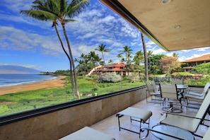 Un balcon donnant sur une plage et des palmiers, un coin repas avec des chaises et une table, et un bâtiment avec un toit rouge en arrière-plan.