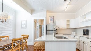 Une cuisine avec des meubles blancs, un îlot central doté d’un évier et un coin repas avec des chaises et une table en bois.