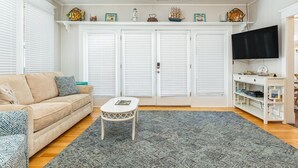 Un salon avec un canapé beige, une petite table blanche, un tapis gris, un ventilateur de plafond et un téléviseur posé sur un meuble blanc.