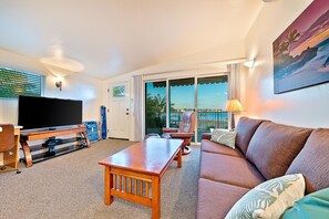 Relax in a spacious room with a flat-screen TV, comfortable sofa, and sliding glass door leading to an outdoor area with a view of palm trees.