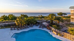 A pool area with lounge chairs, palm trees, and a view of the ocean. - Bay View Villas 203 | Waterfront, Community Pool (Tybee Island)